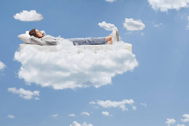 Teenage girl in jeans and hoodie resting on a mattress on clouds up in the sky
