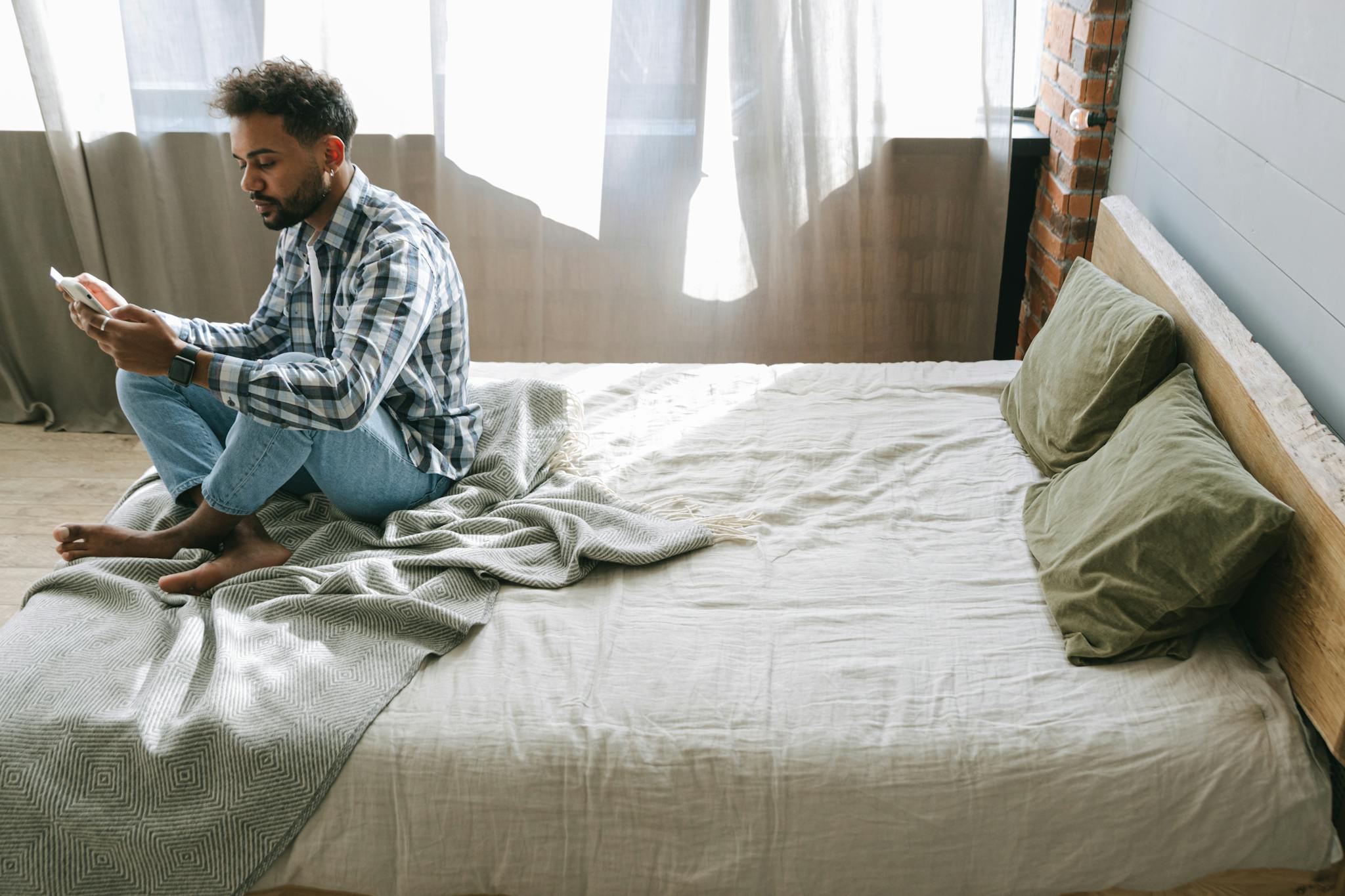 Man in casual attire sitting on bed, using a smartphone in a cozy bedroom.